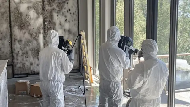 Two inspectors in white protective suits documenting a mold-damaged room with cameras